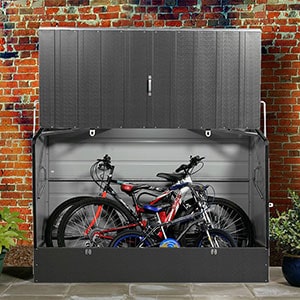 The 6x3 Trimetals Anthracite Protect-a-Cycle Secure Garden Bike Storage, situated on paving slabs, back against a brick wall, and with its lid up to reveal 3 bikes.